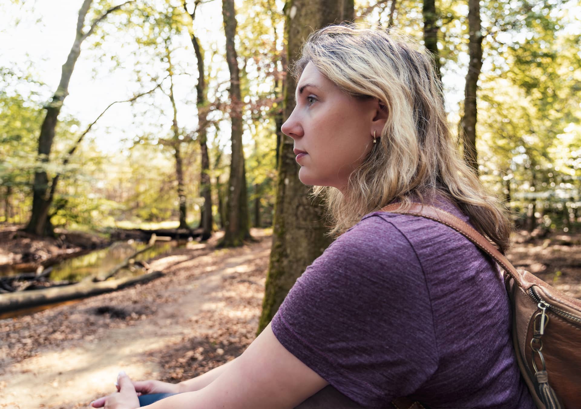 Side profile of a woman with blonde hair wearing a purple shirt, in a sunlit forest with a brown backpack strap visible.