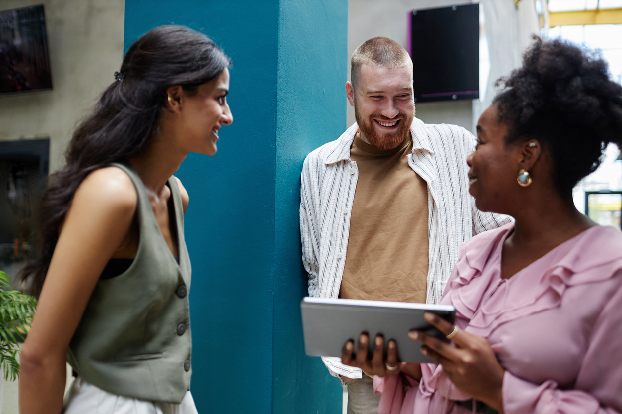 Three coworkers smile and chat near a teal pillar, one holding a tablet.