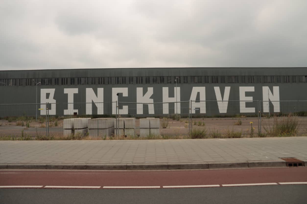 Wide view of a gray industrial warehouse with a giant white 'BINCKHAVEN' sign on its side, a chain-link fence, and a paved sidewalk in front.
