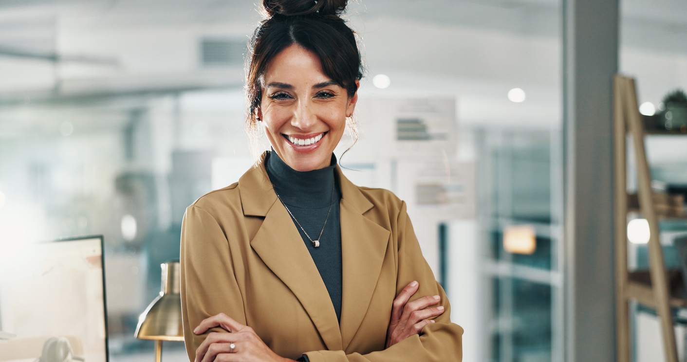 Smiling woman in a tan blazer crosses her arms in a modern office setting.