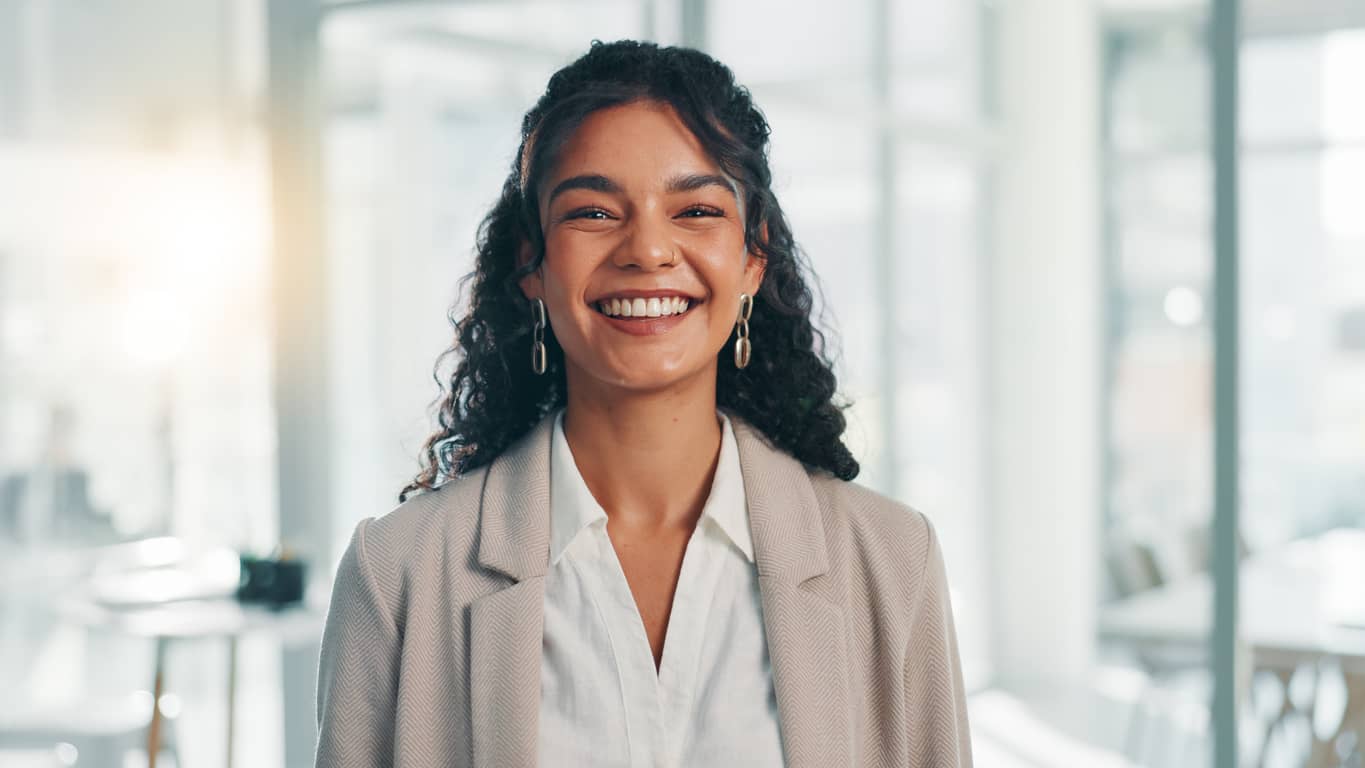 Smiling woman with curly dark hair wearing a beige blazer in a bright office setting.