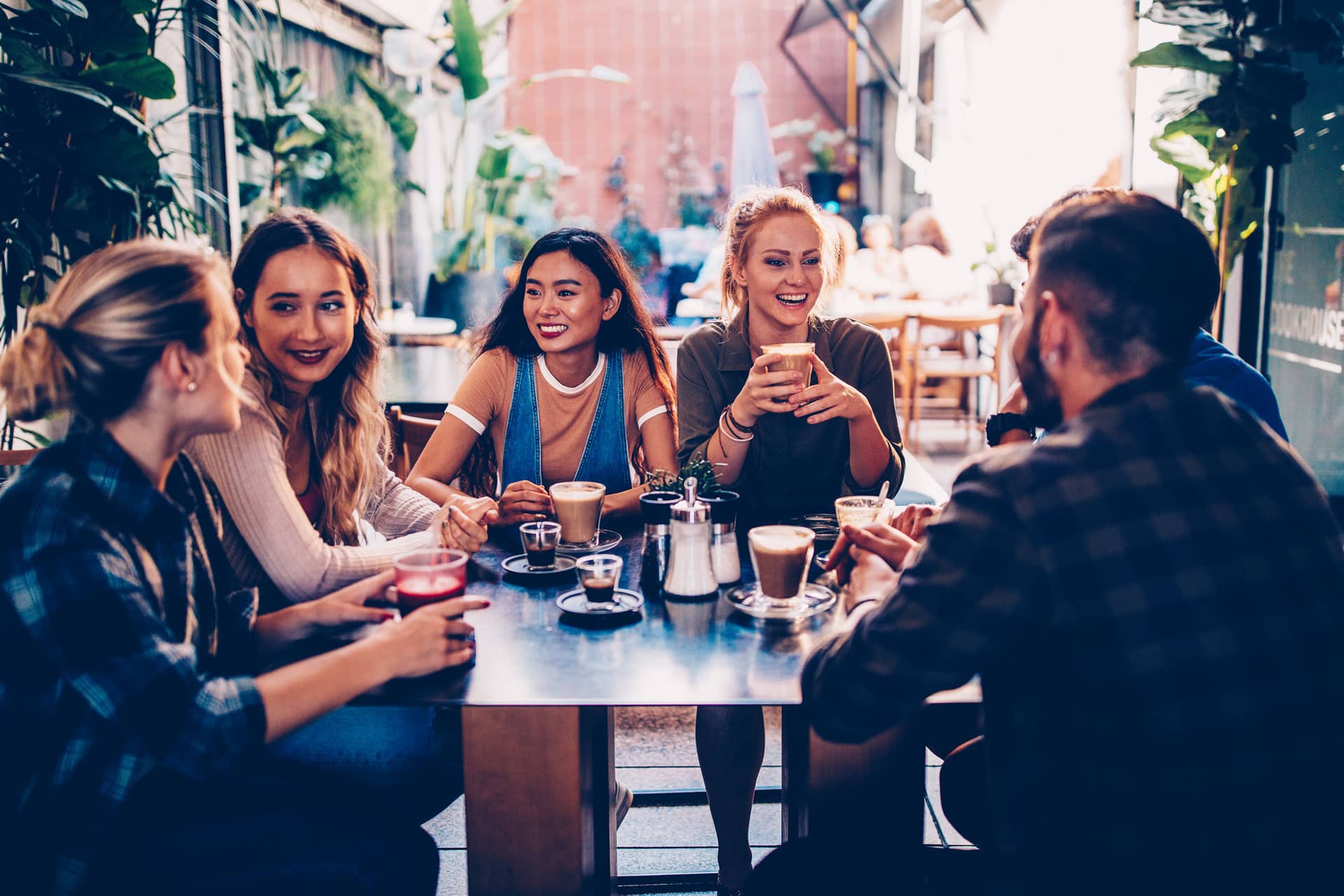 Group of friends sitting at a cafe table, smiling and chatting with coffee drinks in front of them on a sunny outdoor patio.
