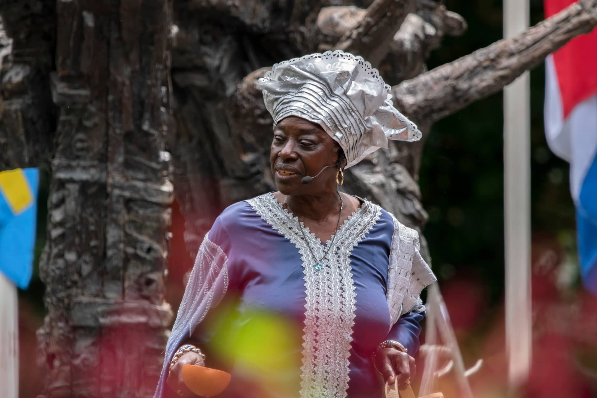 Woman in a purple traditional dress with a silver headwrap, wearing a headset mic at an outdoor cultural event.