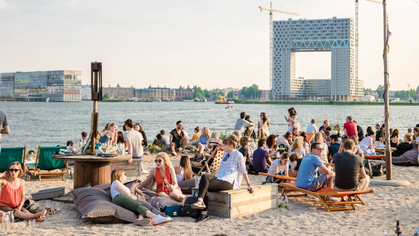 Crowded riverside beach scene with people lounging on bean bags and picnic benches, sunny day by the water with a city skyline in the background.