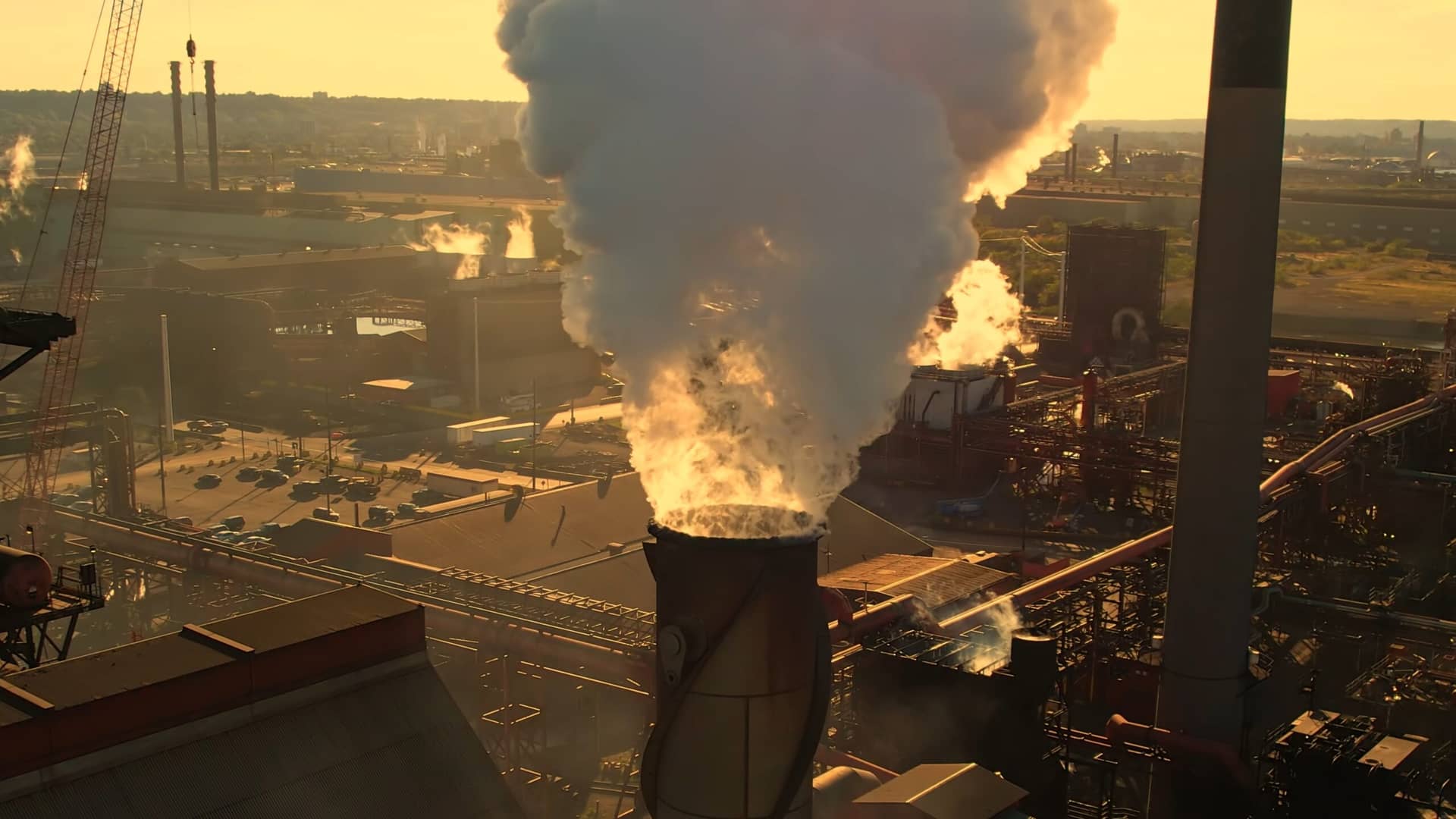 Industrial plant at sunset releasing a thick plume of white steam from a tall smokestack, with cranes and piping in the foreground.