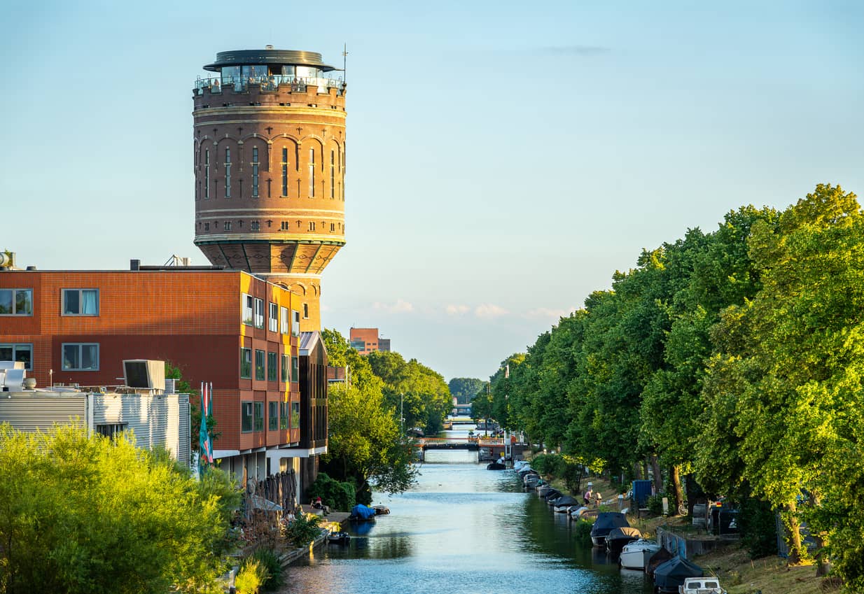 Brick water tower beside a red-orange apartment building overlooking a canal with moored boats and green riverbanks.] , but ensure concise: