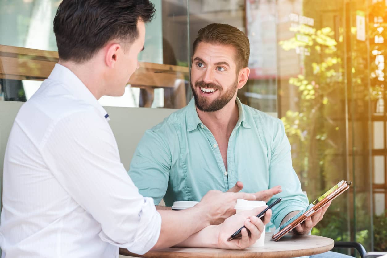 Two men sit at a cafe table sharing a tablet and coffee, engaged in conversation outside a glass-walled space.