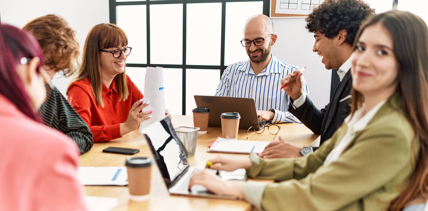 Six coworkers around a wooden conference table, laptops open, papers and coffee cups, smiling and collaborating.
