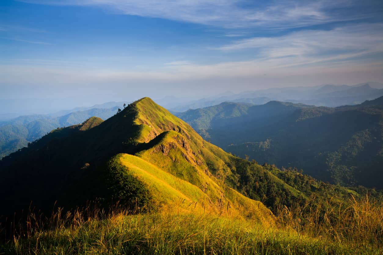 Sunlit green mountain ridge with rolling hills under a blue sky and distant hazy mountains on the horizon.