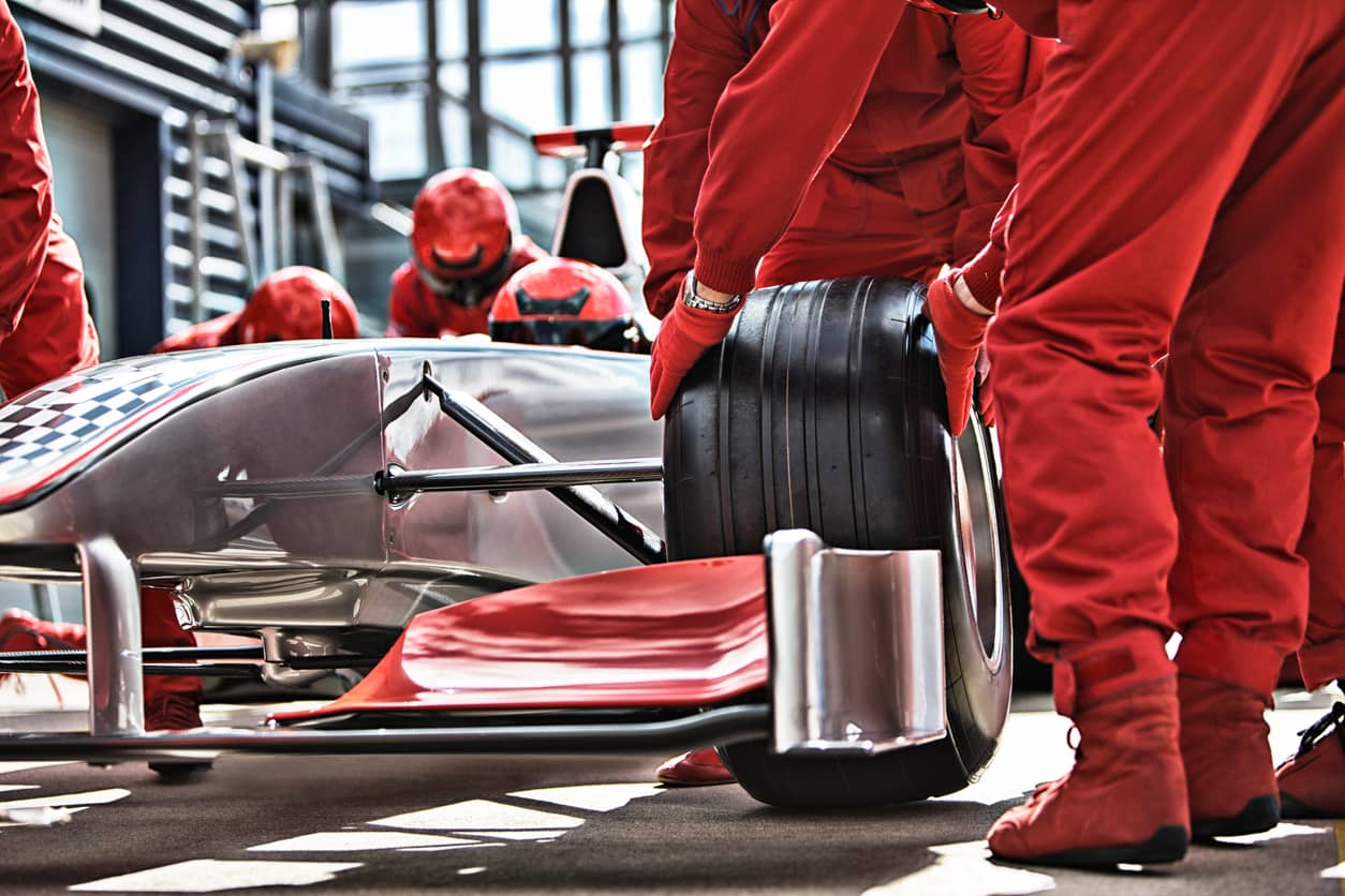Pit crew in red suits changing a tire on a silver race car in the pit lane.
