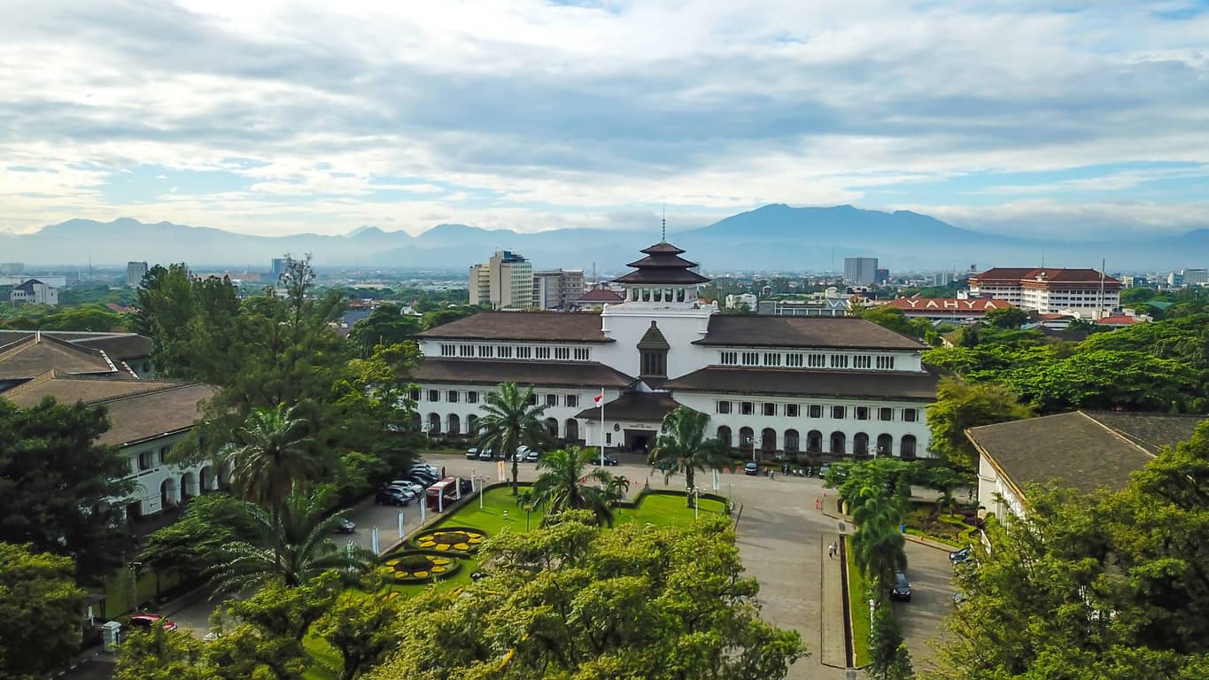 Aerial view of a white, multi-wing building with a tiered central roof and pagoda-like tower, surrounded by palm trees and a landscaped garden, with mountains in the distance.