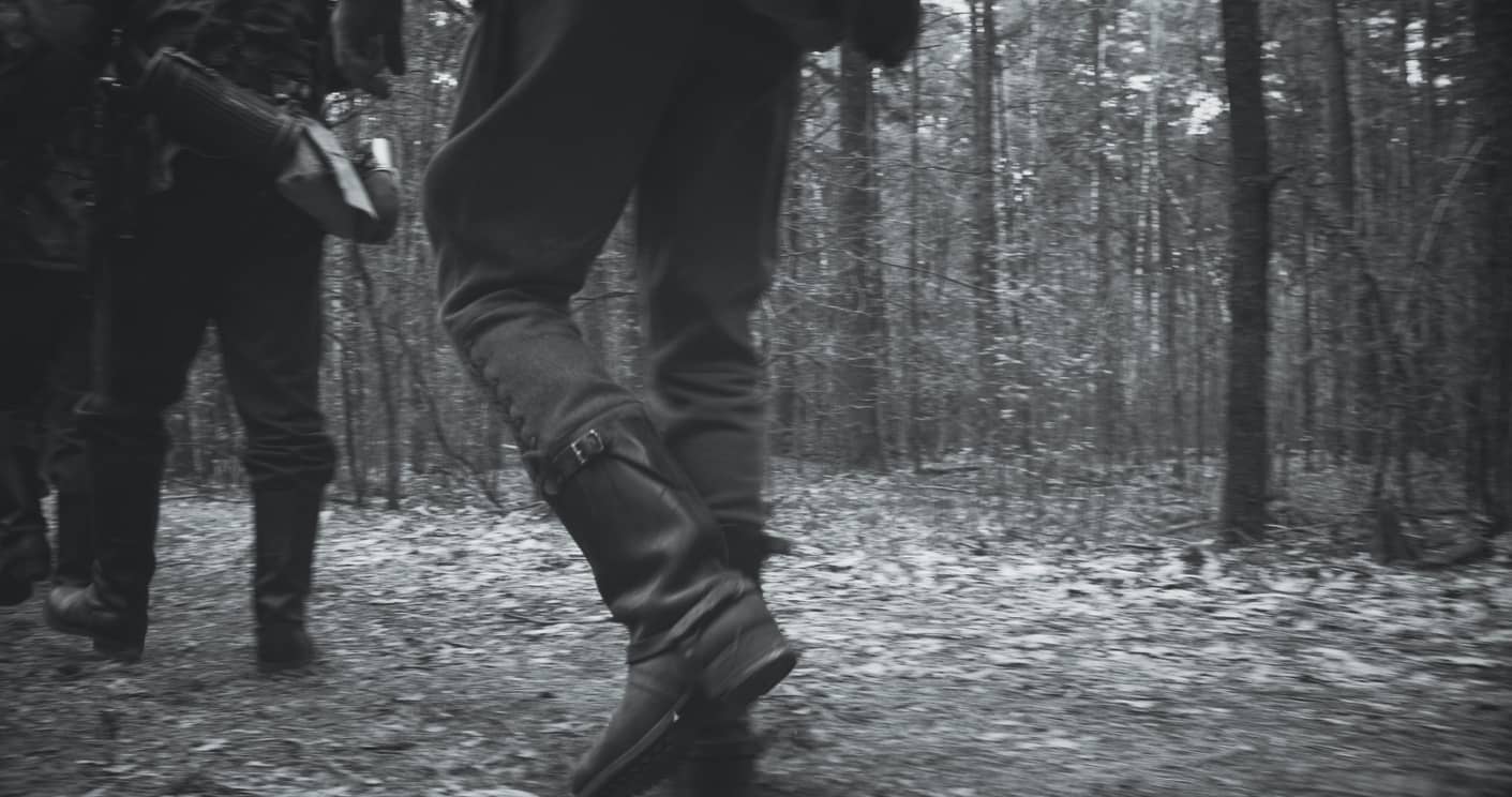 People walking through a leaf-covered forest path; close-up on sturdy boots stepping forward (black-and-white photo).