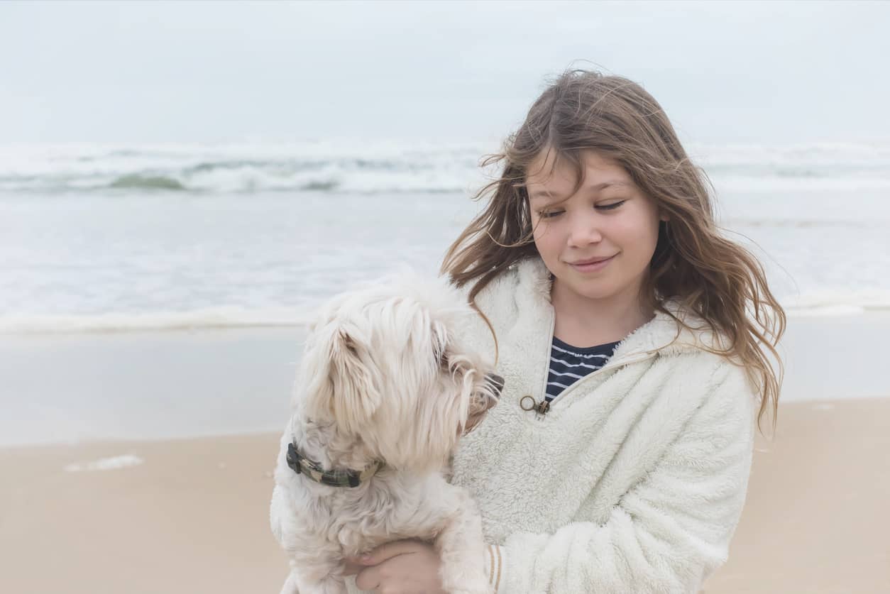 Girl in a white fluffy jacket holding a small white dog at the beach, with waves in the background and a gentle smile on her face.