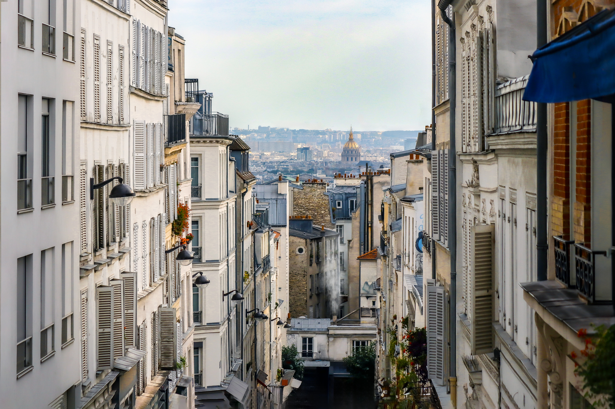 Narrow Parisian street flanked by white townhouses with shuttered windows, leading to a distant city skyline and a dome-topped building.