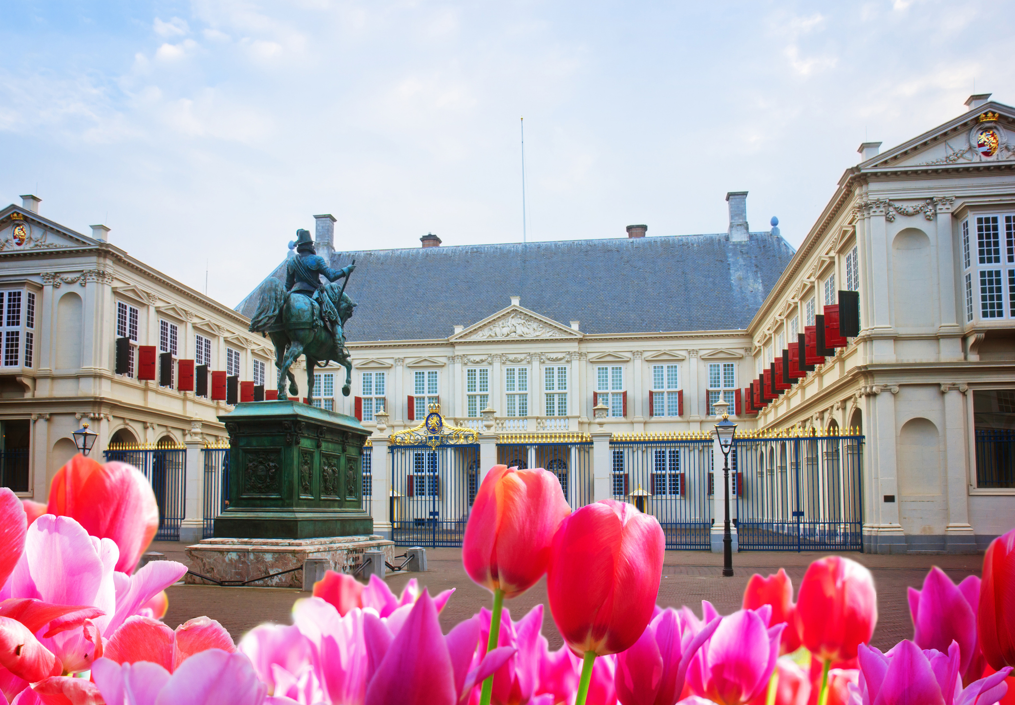 Bronze equestrian statue in a courtyard, with a grand cream-colored neoclassical building and bright pink tulips in the foreground