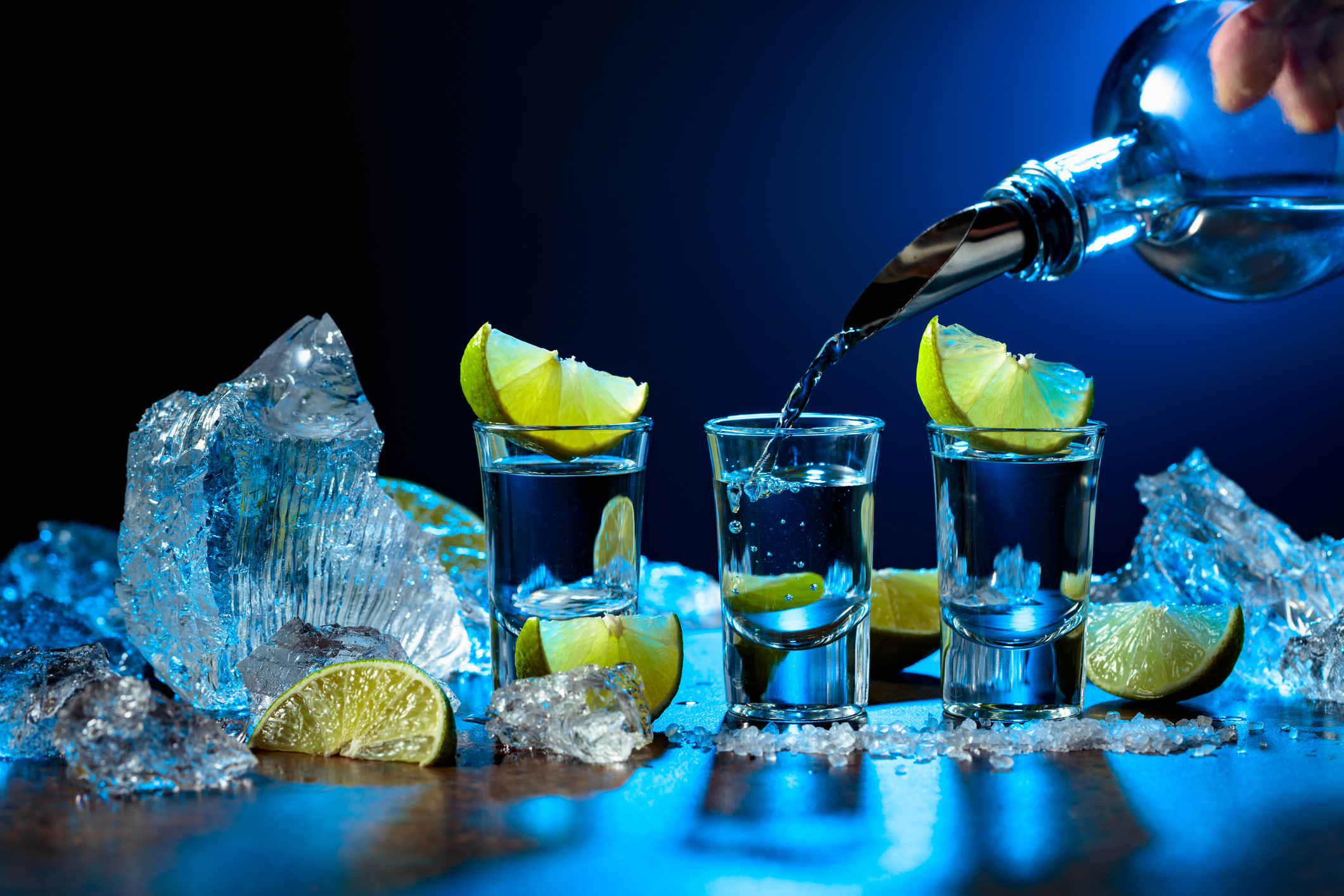 Pouring blue cocktail into shot glasses surrounded by lime wedges and ice with a dark background.