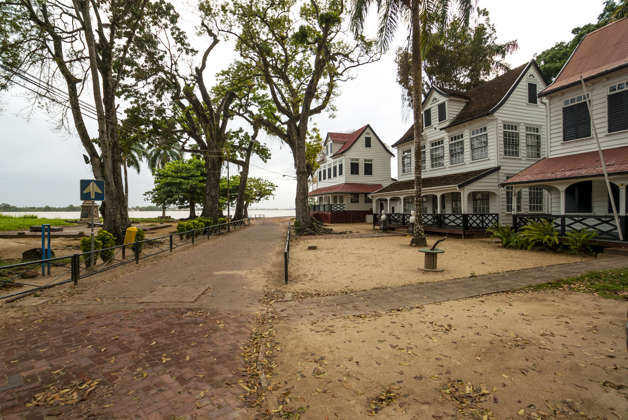 Seaside street with white wooden houses on the right, large trees overhead, a brick pedestrian path, and a blue sign on the left.