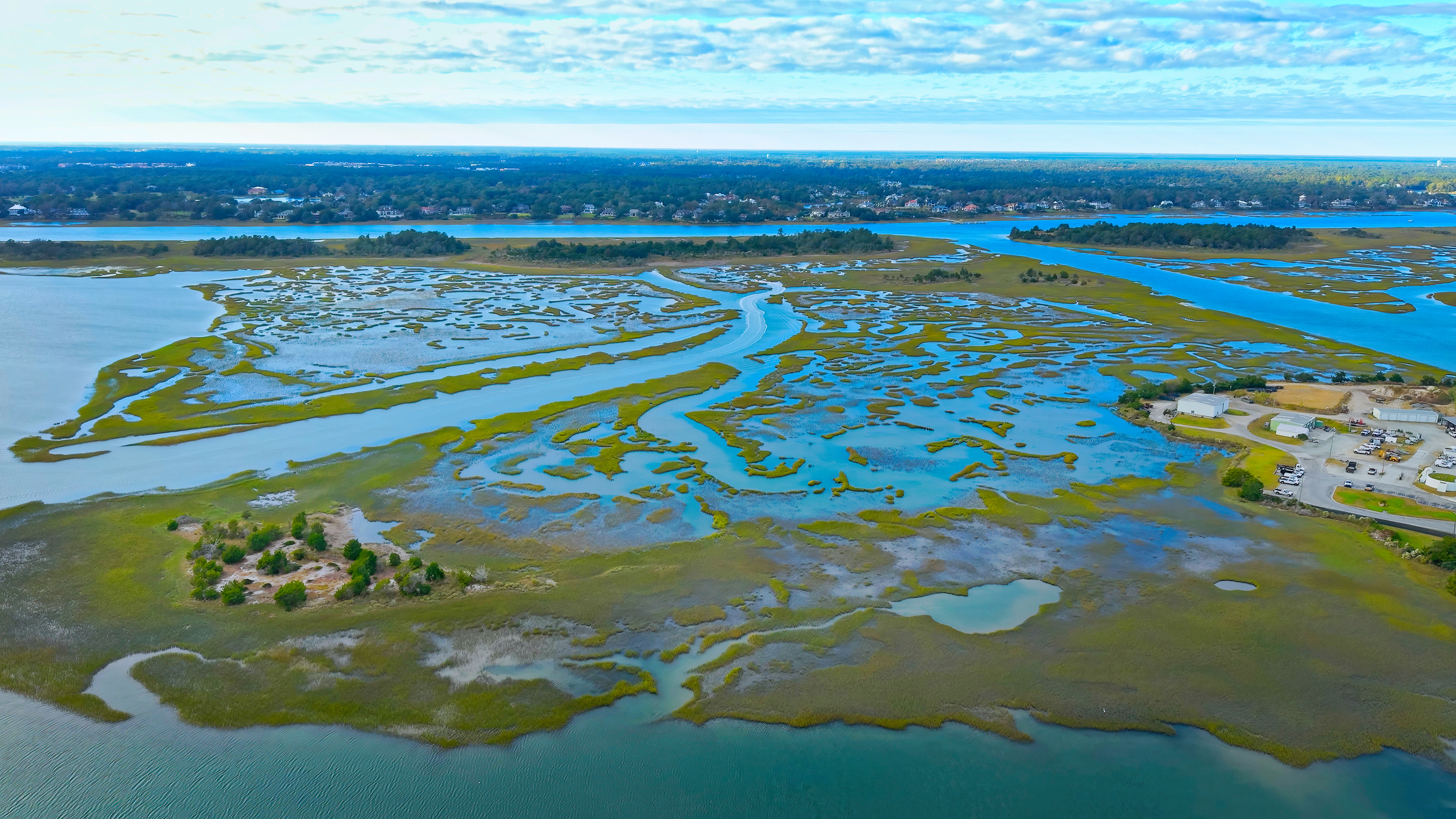 Aerial view of a braided tidal marsh with winding blue waterways and green grassy channels extending toward a distant residential shoreline under a cloudy sky.