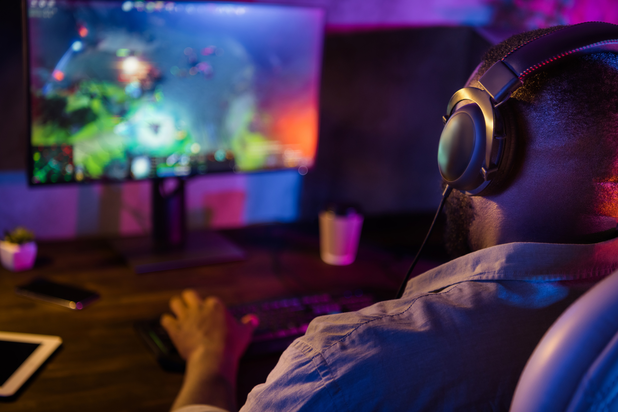 Person gaming at a desk with neon lighting: a blurred monitor displaying a colorful game, a large headset hanging on a chair, and a hand reaching toward the desk.