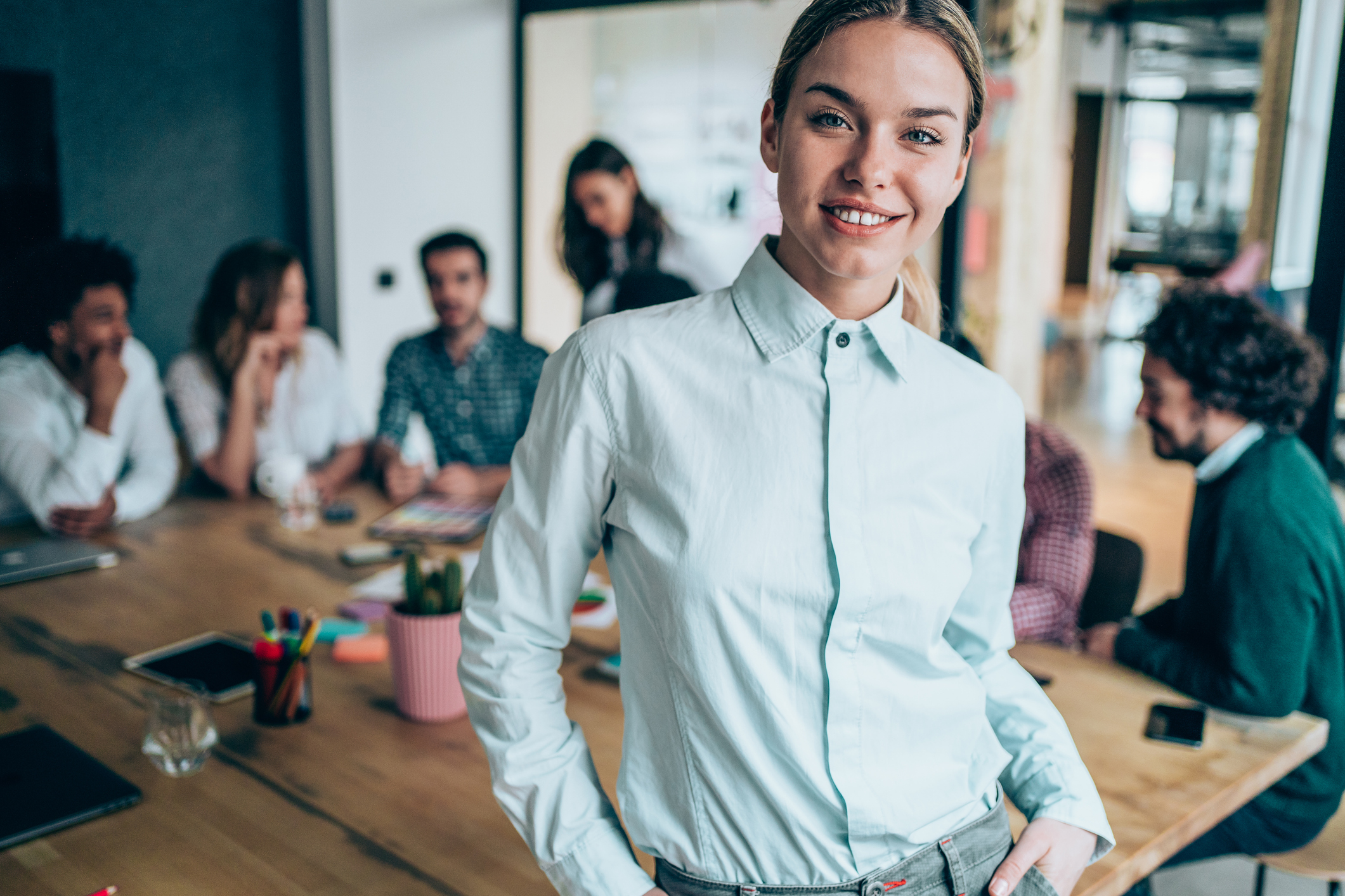 Smiling woman in a light blue shirt stands at an office table, with colleagues blurred in the background.
