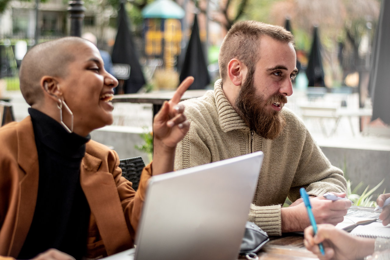 Two colleagues sit at an outdoor cafe, smiling as the woman points and the bearded man reviews papers and a laptop.