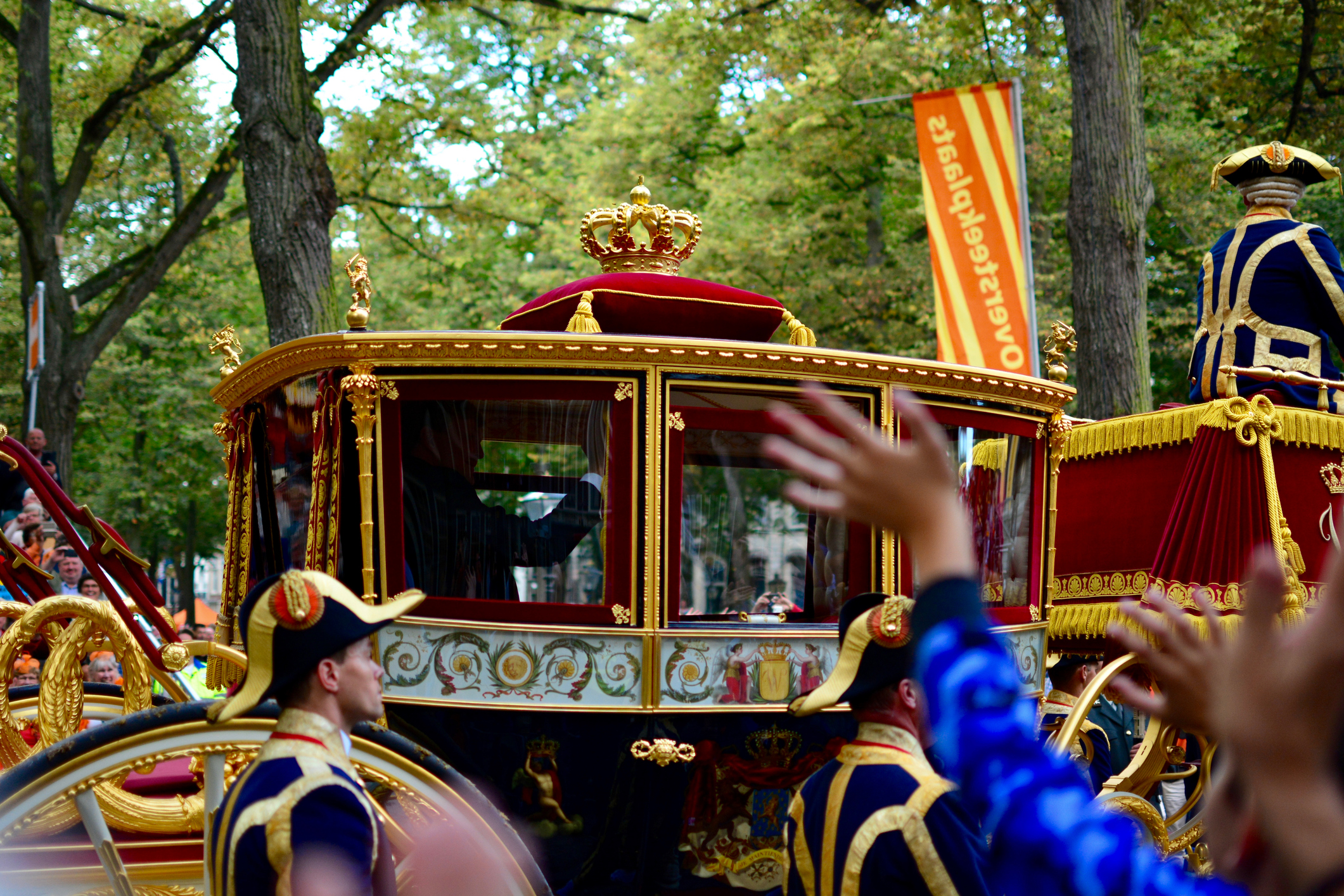 Ornate gold carriage with red velvet, crowned top, in a royal parade among spectators and uniformed guards in blue and gold.",