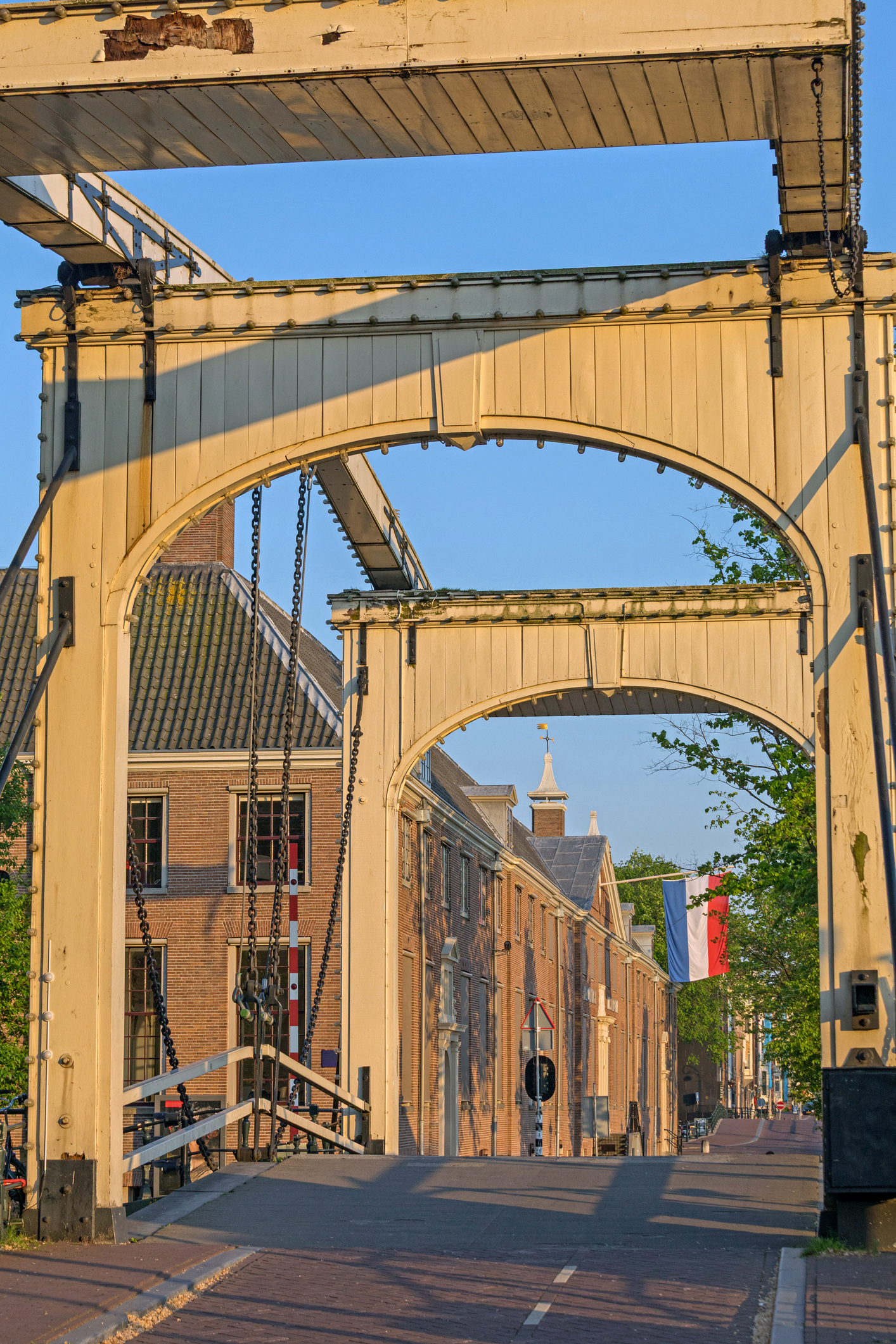 Cream-colored arched drawbridge structure over a brick street, with hanging chains and adjacent brick buildings under a blue sky; a French flag hangs to the right.