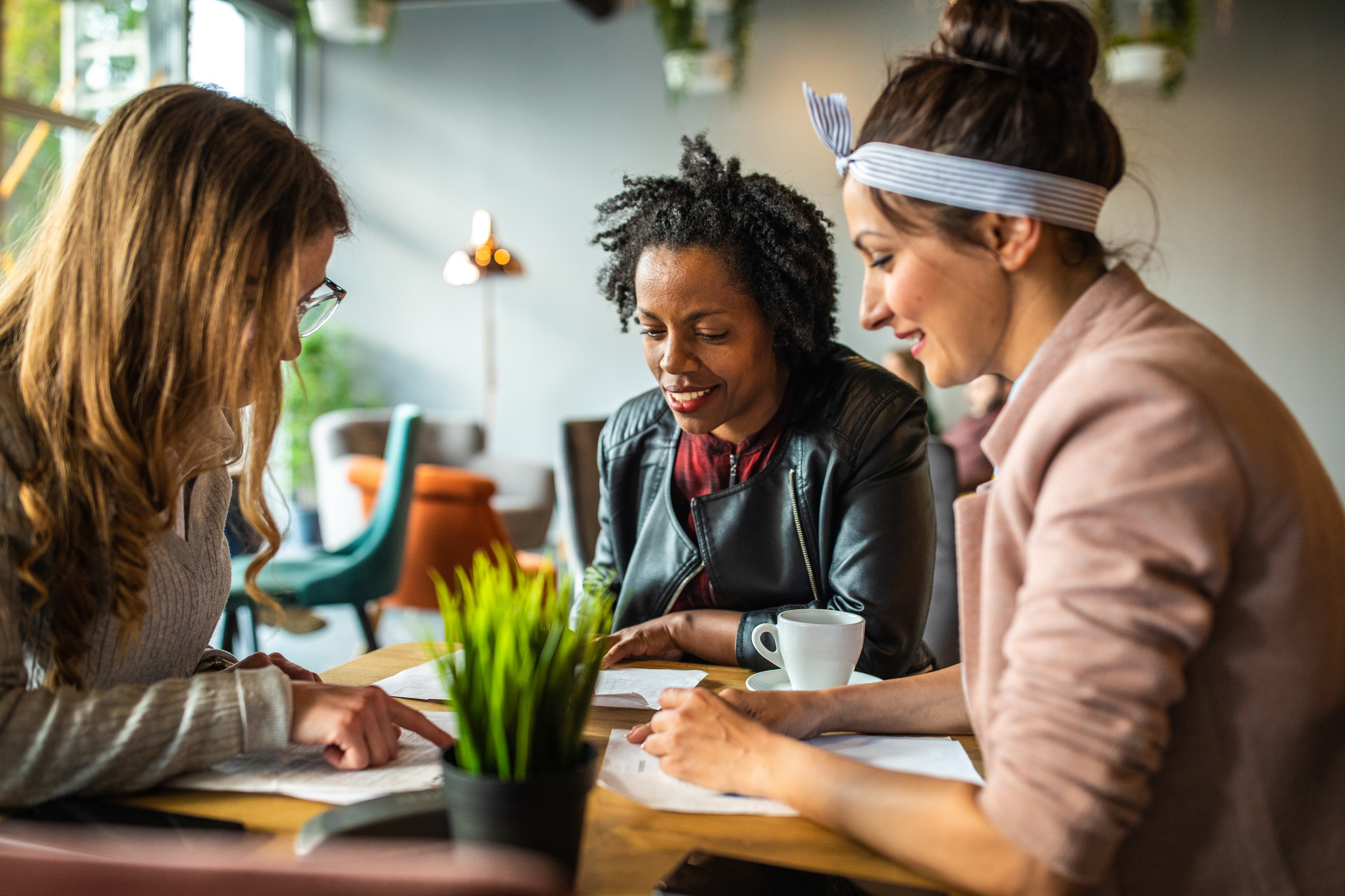 Three women sit at a café table, looking over documents together with a small plant in the foreground and coffee cups nearby.