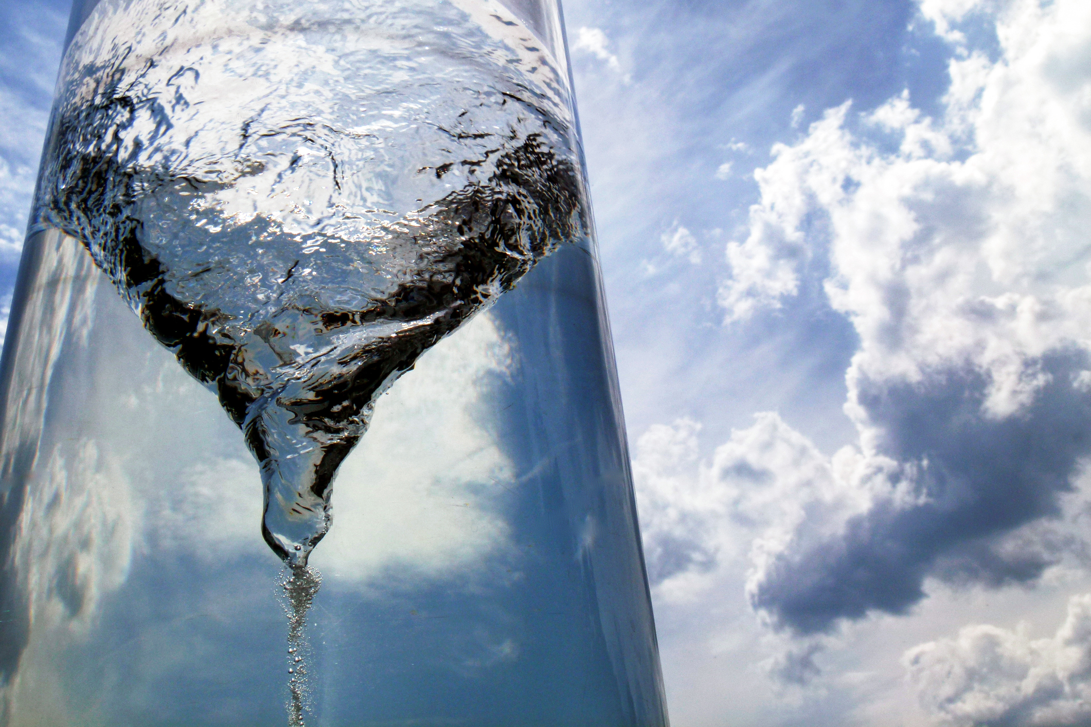 Close-up of a water droplet sliding down a curved glass surface with a blue sky and white clouds behind it.
